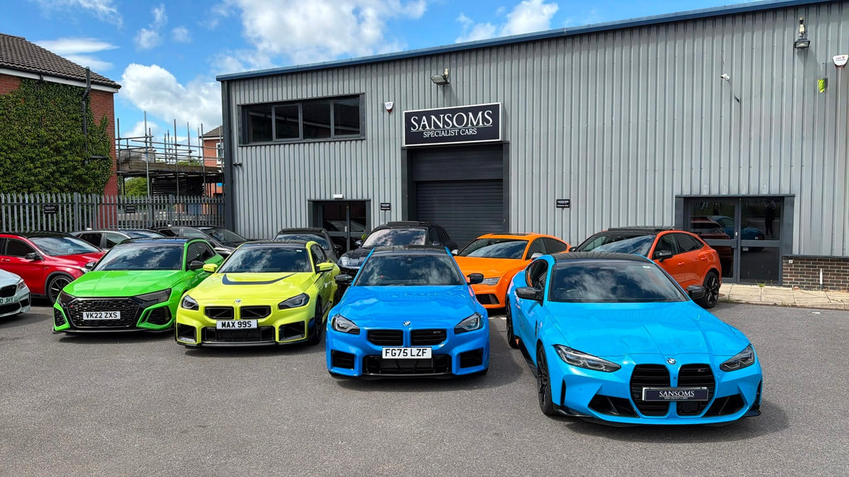 A lineup of five BMW sports cars in vibrant colors (red, green, yellow, blue, and light blue) parked in front of a gray industrial building with 'SANSOMS MOTORS CAR' signage.